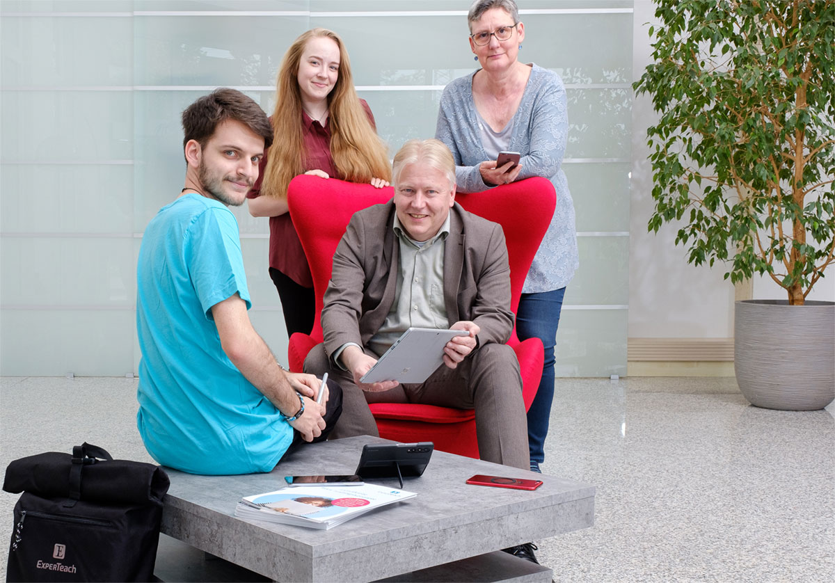 1 Mann sitzt auf rotem Sessel, 2 Frauen stehen dahinter, 1 Mann sitzt davor - Data Center & Cloud Consulting